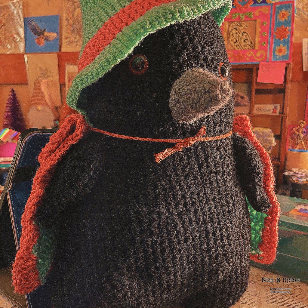 Crocheted crow as a stuffed toy with a hat and cape on in a Wes Anderson style coloring.
