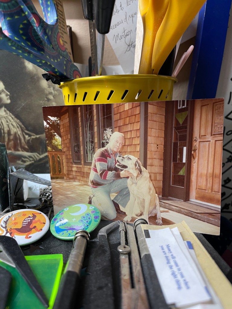 Photo of a man sitting next to a dog that is on a desk with items around it.