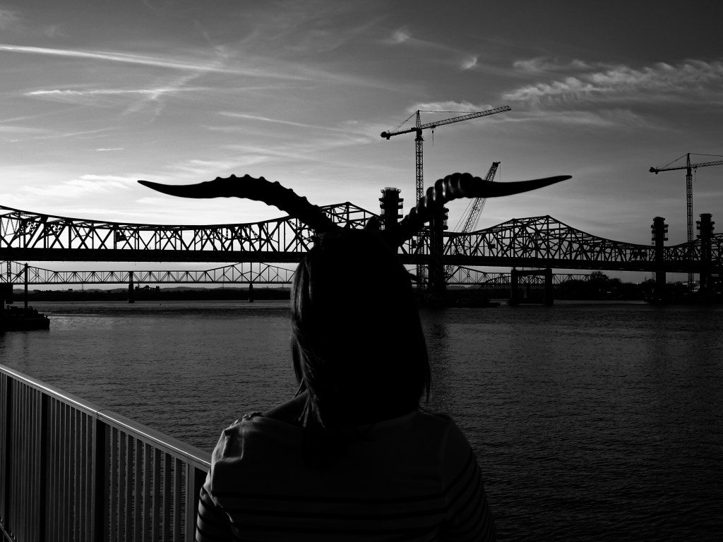 A black and white photograph of a woman wearing horns staring over a river at a pair of bridge.